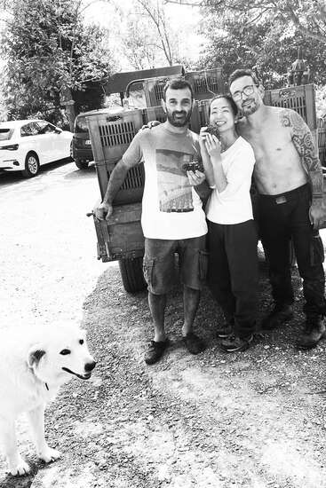 Three people pose happily outdoors by a truck, one holding grapes, another shirtless with tattoos. A white dog stands in the foreground, and cars are parked behind.