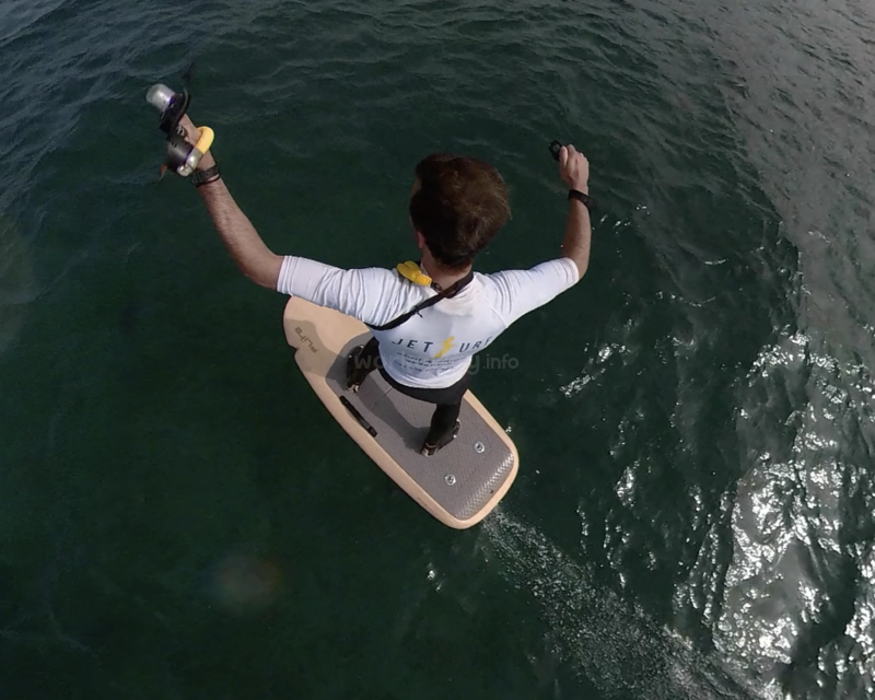 The image depicts a man standing on a board in the water, with his arms raised and holding an object, likely engaged in water sports or activities.