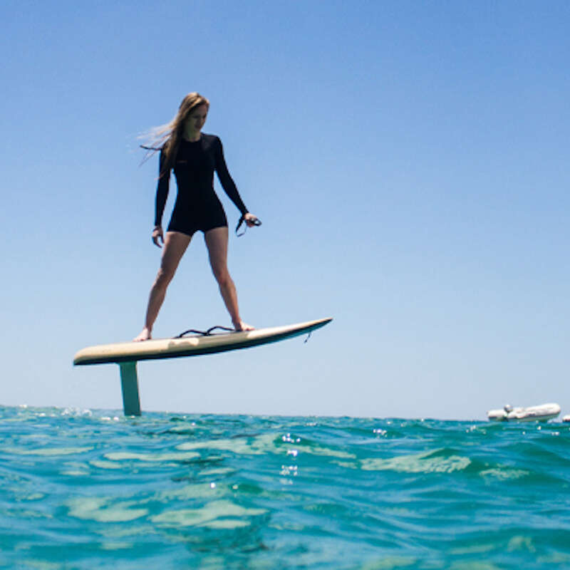 The image depicts a woman standing on a hydrofoil board in the ocean, wearing a black wetsuit, with the board\'s foil extending below the water\'s surface.