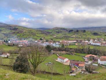 A picturesque rural village nestles among rolling green hills, scattered houses with red rooftops, trees, and pastures under a dramatic, cloudy sky, creating a serene landscape.