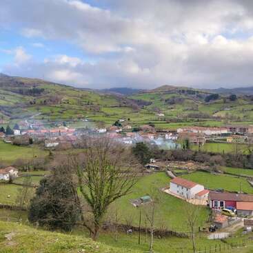 Ein malerisches, ländliches Dorf schmiegt sich an sanfte, grüne Hügel, verstreute Häuser mit roten Dächern, Bäume und Weiden unter einem dramatischen, wolkenverhangenen Himmel, der eine heitere Landschaft schafft.