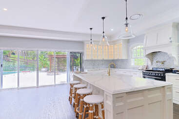This kitchen features a large marble island with four stools, pendant lights, white cabinetry, a modern stove, and sliding glass doors opening to a sunny patio.