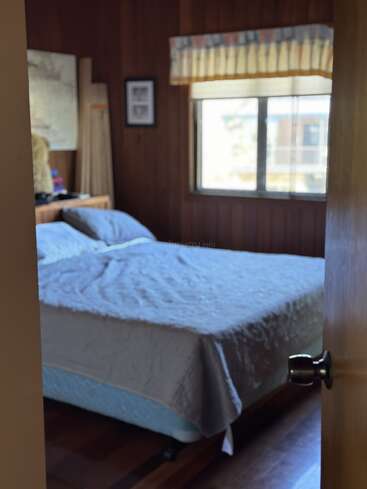 This cozy bedroom has wood-paneled walls, a neatly made bed with a gray blanket, a large window with a valance, and soft natural light shining in.