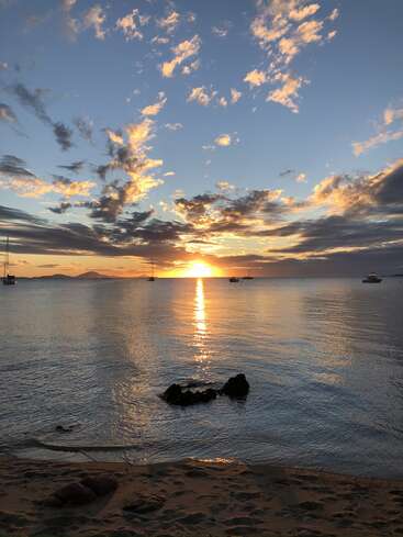 Golden sunlight reflects across calm ocean water at sunset, with scattered clouds, anchored sailboats, rocky shoreline, sandy beach, and distant land silhouettes beneath a tranquil sky.
