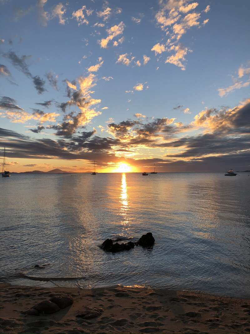 Golden sunlight reflects across calm ocean water at sunset, with scattered clouds, anchored sailboats, rocky shoreline, sandy beach, and distant land silhouettes beneath a tranquil sky.