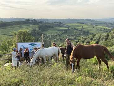 Um grupo de pessoas com crianças e dois cavalos se reúnem ao ar livre em um grande quadro, cercado por pitorescas colinas verdes sob um céu claro.