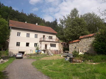 Une maison à deux étages avec un toit rouge se trouve à côté d'un bâtiment rustique en pierre, entourée d'une végétation luxuriante, de voitures garées et d'un ciel partiellement nuageux.