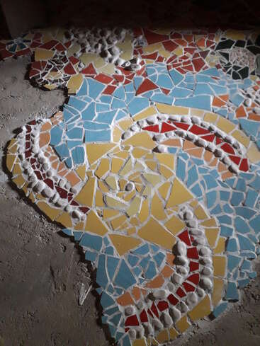 Cette image montre une mosaïque colorée sur le sol, composée de carreaux brisés de couleur rouge, jaune, bleue et blanche, formant des motifs géométriques abstraits et des courbes.