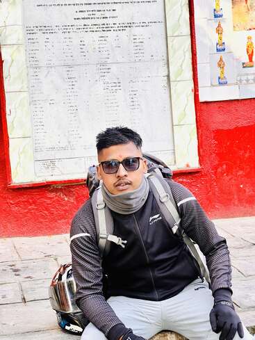 Um homem usando óculos escuros, luvas e uma mochila senta-se no chão perto de um capacete. Atrás dele há uma parede vermelha com pôsteres e um quadro branco.