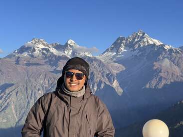 Uma pessoa sorridente, usando óculos escuros e roupas de inverno, fica ao ar livre com impressionantes picos de montanhas cobertas de neve e céu azul claro no fundo cênico. Vista bonita e tranquila.