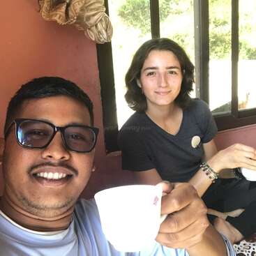 Duas pessoas sentadas juntas, sorrindo e saboreando bebidas quentes em xícaras brancas. Elas estão em um ambiente interno, perto de uma janela com luz natural, parecendo relaxadas e felizes.