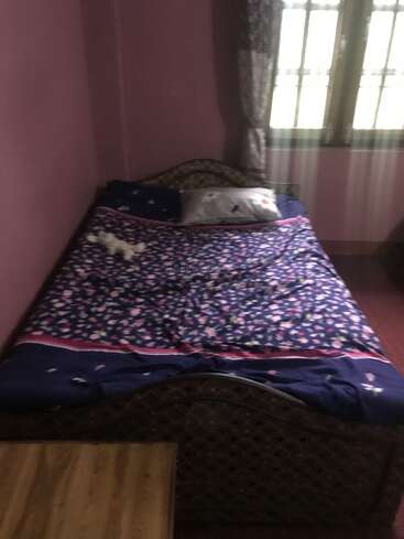 Esta imagem mostra um quarto aconchegante com uma cama de madeira, lençol floral, travesseiros, paredes cor-de-rosa, uma janela com cortinas e uma pequena mesa de madeira ao lado da cama.