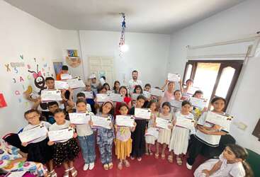 Dans une salle de classe, un groupe d'enfants souriants tient fièrement des certificats. Deux adultes se tiennent derrière eux. La salle de classe est décorée de lettres alphabétiques colorées.