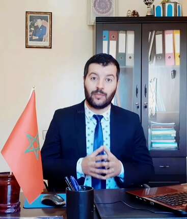Un homme en costume est assis à un bureau derrière lequel se trouvent un drapeau marocain, des fournitures de bureau et des dossiers. Il semble expliquer ou parler de quelque chose.