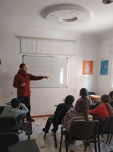 A teacher stands near a whiteboard, pointing and engaging with a group of attentive students seated in a classroom. Educational posters decorate the white walls.