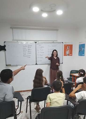 A teacher stands by a whiteboard teaching English words and numbers to engaged students in a brightly lit classroom; several children eagerly raise their hands.
