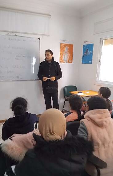 A teacher stands in front of a whiteboard, teaching a group of attentive students in a classroom. Posters are on the wall, sunlight enters.