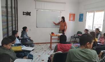 A teacher stands at a whiteboard, engaging a group of students seated in a classroom. The students listen attentively, some holding notebooks and worksheets.