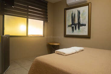 A cozy bedroom with beige walls features a neatly made bed, abstract painting, window with sunset view, mini fridge, wall-mounted TV, and air conditioner.