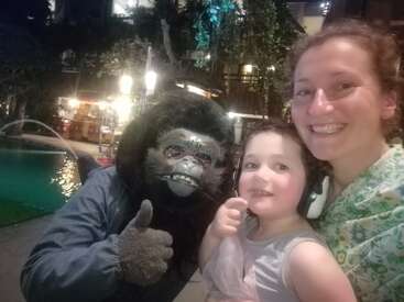 Una mujer y un niño sonríen para un selfie con una persona disfrazada de gorila que les hace un pulgar hacia arriba, cerca de una piscina por la noche, fondo iluminado.