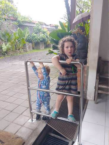 Dos niños pequeños se suben a una barandilla metálica en una entrada exterior. Detrás de ellos hay exuberantes plantas verdes, una estatua tallada y un patio de baldosas.