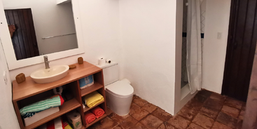 Cette salle de bains comprend un lavabo avec un comptoir en bois, des étagères ouvertes avec des serviettes colorées, des toilettes, une douche, un miroir et un sol carrelé rustique.