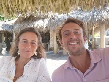 Une femme et un homme sourient ensemble dans un cadre tropical en plein air sous un toit de chaume, profitant d'une journée ensoleillée dans une ambiance de vacances détendue et un décor naturel.