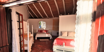 Cette chambre confortable et ensoleillée comprend deux lits, du carrelage en terre cuite, des poutres en bois apparentes, des murs blancs, des rideaux en dentelle et un décor rustique, créant ainsi une atmosphère chaleureuse et accueillante.