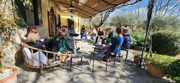 A group of people sit in a circle on a sunny terrace, surrounded by plants and trees, engaging in conversation under a rustic awning next to a house.
