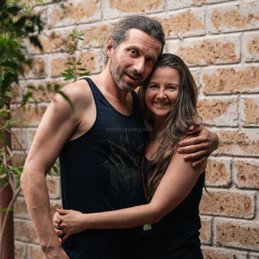 A smiling couple stands closely together, embracing warmly. They wear casual dark clothing and appear happy. The background features a rustic brick wall and green plants.