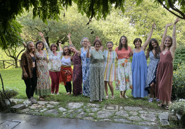 A joyful group of thirteen women stands together outdoors, smiling and raising their hands, surrounded by lush green trees and plants on a sunny day.