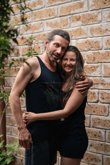 A smiling couple stands closely together, embracing warmly. They wear casual dark clothing and appear happy. The background features a rustic brick wall and green plants.