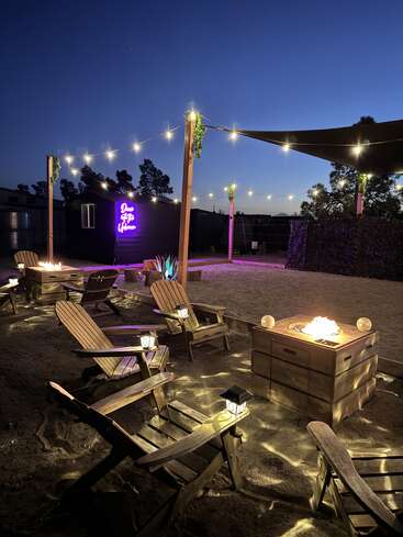 Cadeiras de madeira circundam uma aconchegante fogueira em um terreno arenoso, iluminada por luzes de cordas no alto. Um letreiro de neon em uma parede escura diz "Desert Life Lifewow".