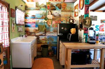 Das Bild zeigt eine kompakte Küche mit Spüle, Kühlschrank und Kaffeemaschine, mit einer Collage aus Gemälden an der Wand und einem Holztisch mit einem Mixer.