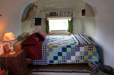 Das Bild zeigt ein gemütliches Schlafzimmer mit Bett, Nachttisch und Lampe, mit einer Patchworkdecke und einem Fenster mit grünen Vorhängen, vor einer weißen Wand.