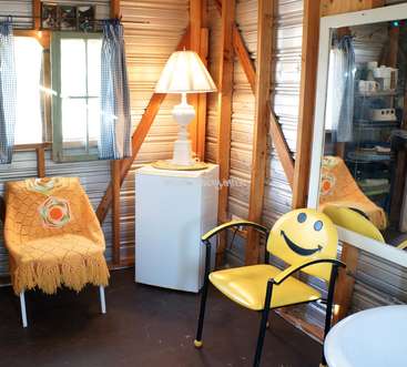 Das Bild zeigt ein gemütliches Zimmer mit einer weißen Lampe, einem gelben Stuhl mit einem Smiley-Gesicht und einem weißen Kühlschrank, umgeben von Holzbalken und einem Fenster.