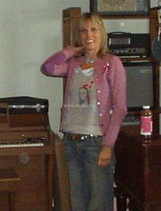 Das Bild zeigt eine Frau, die in einem Raum mit einem Plattenspieler und einem Regal mit Vintage-Elektronik steht und eine rosa Strickjacke und Jeans trägt.
