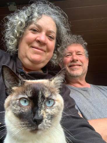 Ein lächelndes Paar mit lockigem, grauem Haar posiert für ein Selfie. Im Vordergrund schaut eine auffällige Siamkatze mit blauen Augen direkt in die Kamera.