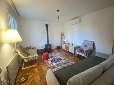Dieses gemütliche Wohnzimmer verfügt über einen Holzofen, einen gemusterten Teppich, bequeme Sitzgelegenheiten, stimmungsvolle Beleuchtung, eine minimalistische Einrichtung, Holzböden und eine ruhige, einladende Atmosphäre mit viel Tageslicht.