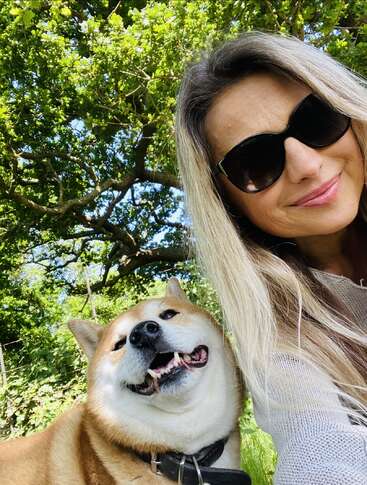A smiling woman wearing sunglasses takes a selfie outdoors with her happy Shiba Inu dog. Bright green trees and sunlight create a cheerful, nature-filled background.