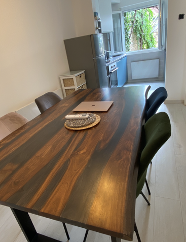 The image shows a modern kitchen-dining area with a wooden table, four chairs, a laptop, phone, and coaster. An open window overlooks greenery outside.
