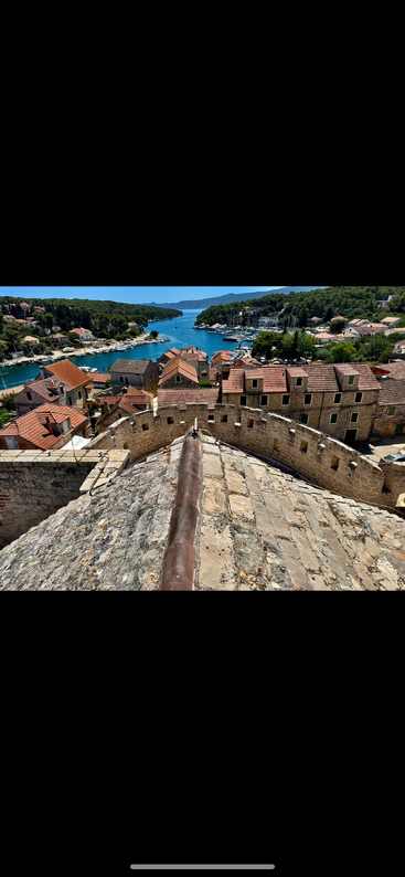 Esta imagen muestra un pintoresco pueblo costero con casas de tejados de terracota, muros históricos de piedra, exuberantes colinas verdes, las aguas azules del puerto, barcos amarrados y un cielo despejado y soleado.