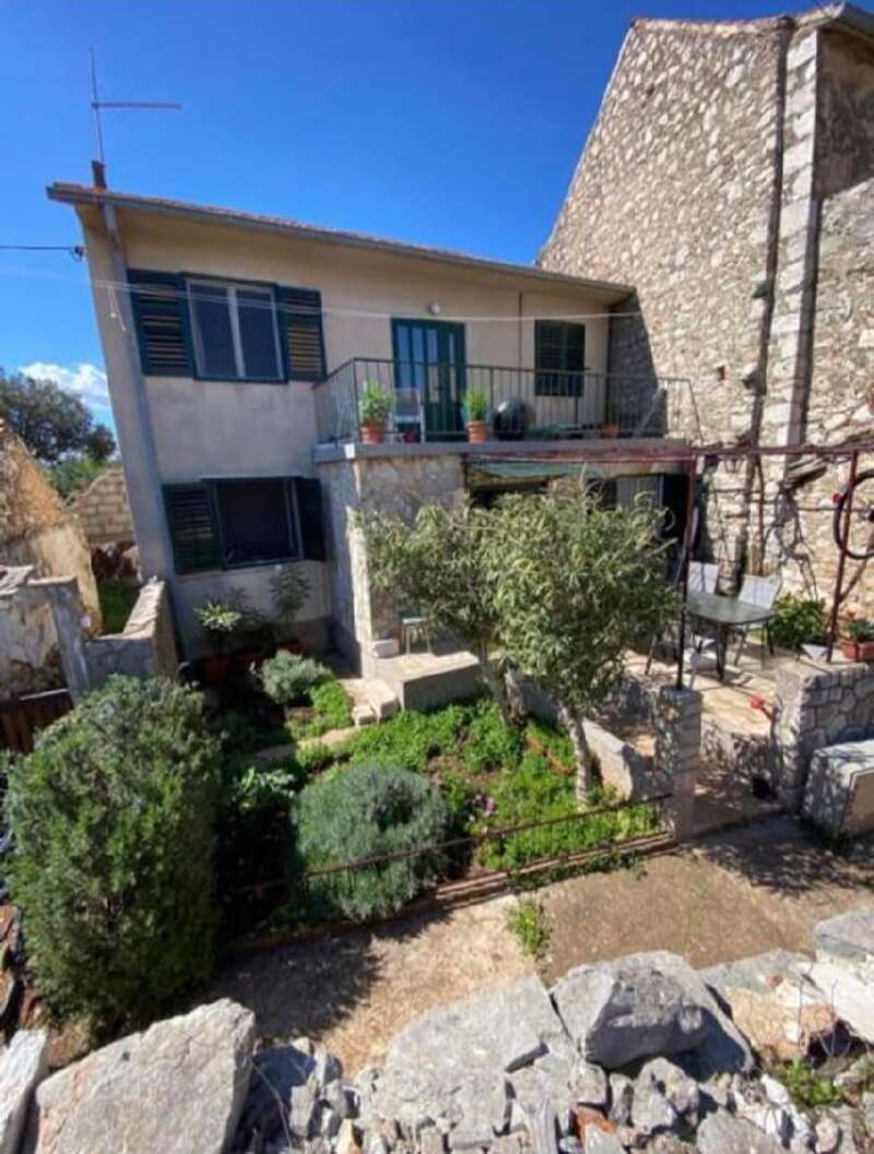 The image depicts a two-story house with a balcony and a garden, featuring stone walls and a serene atmosphere, set against a clear blue sky.