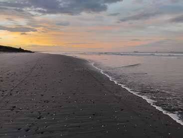 Una playa serena y vacía al atardecer, con suaves olas que se encuentran con arena oscura. El cielo se llena de colores pastel, creando una atmósfera tranquila y sosegada.