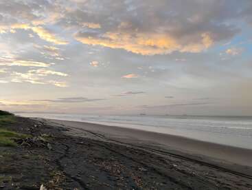Una tranquila playa vacía al amanecer, con arena oscura, trozos de madera esparcidos, suaves olas, un horizonte brumoso y un cielo resplandeciente con suaves nubes naranjas y azules.