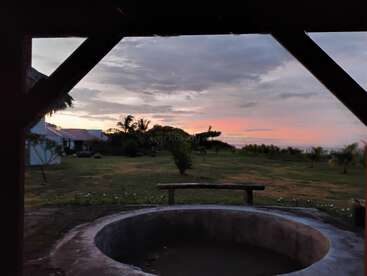 Una estructura circular de hormigón en primer plano conduce a un exuberante patio verde, enmarcado por vigas de madera, contra un colorido cielo al atardecer con nubes dispersas.