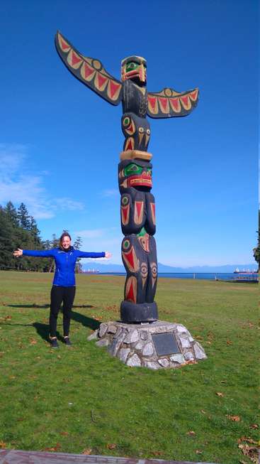 L'image représente une femme debout à côté d'un totem dans un champ herbeux, avec un plan d'eau et des arbres visibles à l'arrière-plan par une journée ensoleillée.