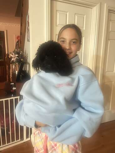 Uma garota sorridente está dentro de casa, vestindo um moletom azul claro. A cabeça de um pequeno cachorro preto espreita de dentro do moletom, criando um momento fofo e engraçado.