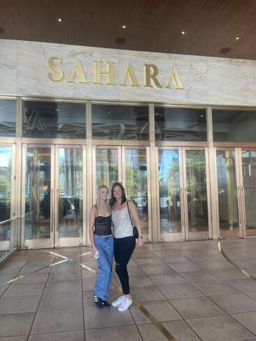 Duas mulheres estão sorrindo em frente à entrada do Saara, com portas de vidro e letras douradas. Elas parecem felizes, vestidas casualmente, desfrutando de um dia agradável juntas.