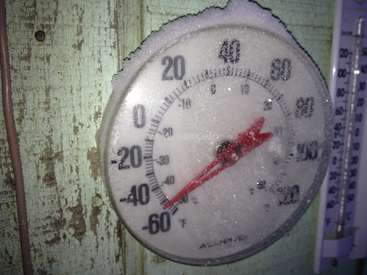 Das Bild zeigt ein rundes Thermometer mit einer roten Nadel, das mit Schnee oder Eis bedeckt ist und an einer Holzwand mit einem Lineal daneben befestigt ist.
